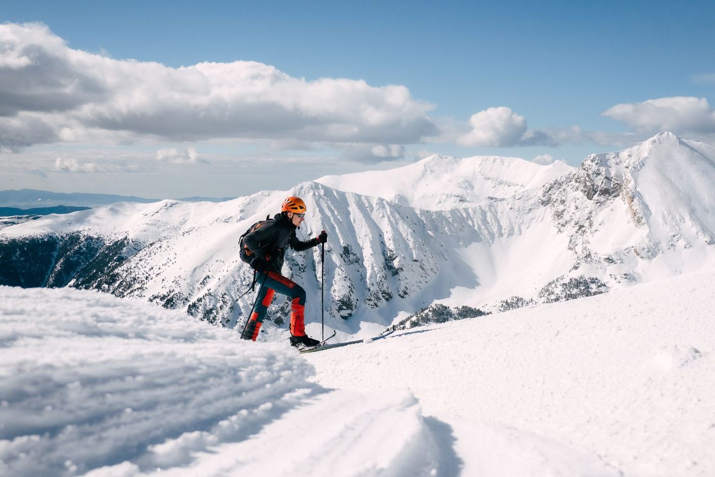 A ski mountaineer climbing a corniced snow ridge with a rucksack, high peaks and cloud below.