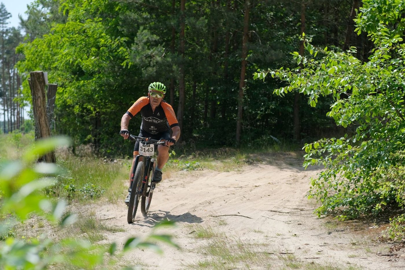 A cross country racer in orange kit riding a fast singletrack line through a sunlit green forest.