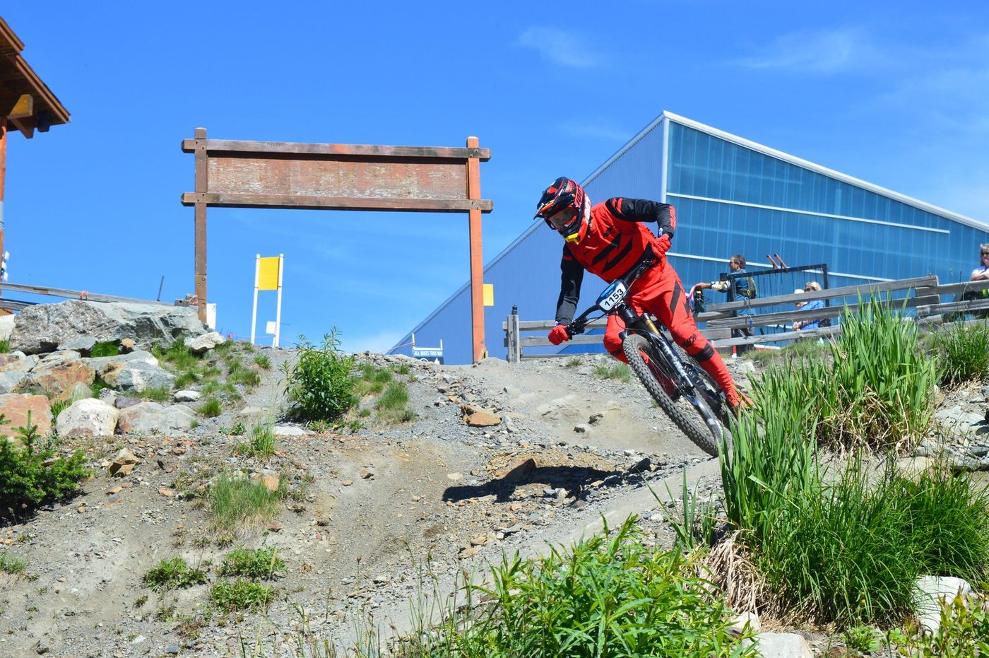 A downhill racer in a red race suit railing a bermed dirt corner at a bike park under a clear blue sky.