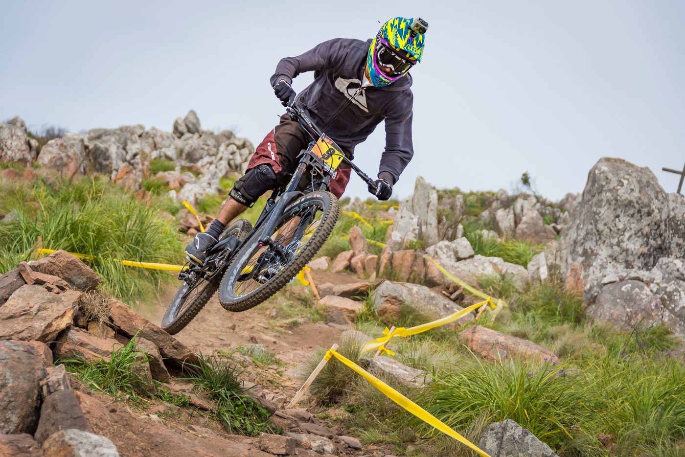 A rider in a full-face helmet dropping into a rocky race line marked with yellow tape, both wheels off the ground.