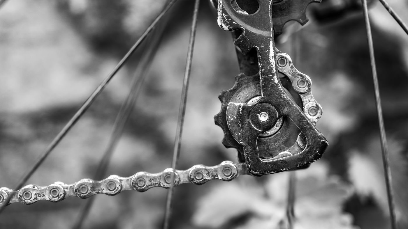 Black and white close-up of a scratched rear derailleur cage and jockey wheel, chain running over worn teeth.