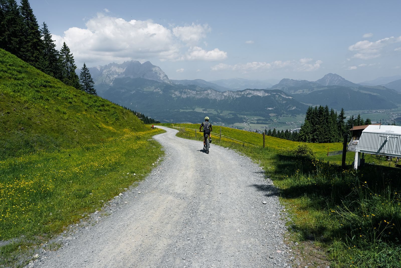 A lone cyclist riding a gravel road that winds through a green alpine pass, with snow-streaked peaks and cloud on the horizon.