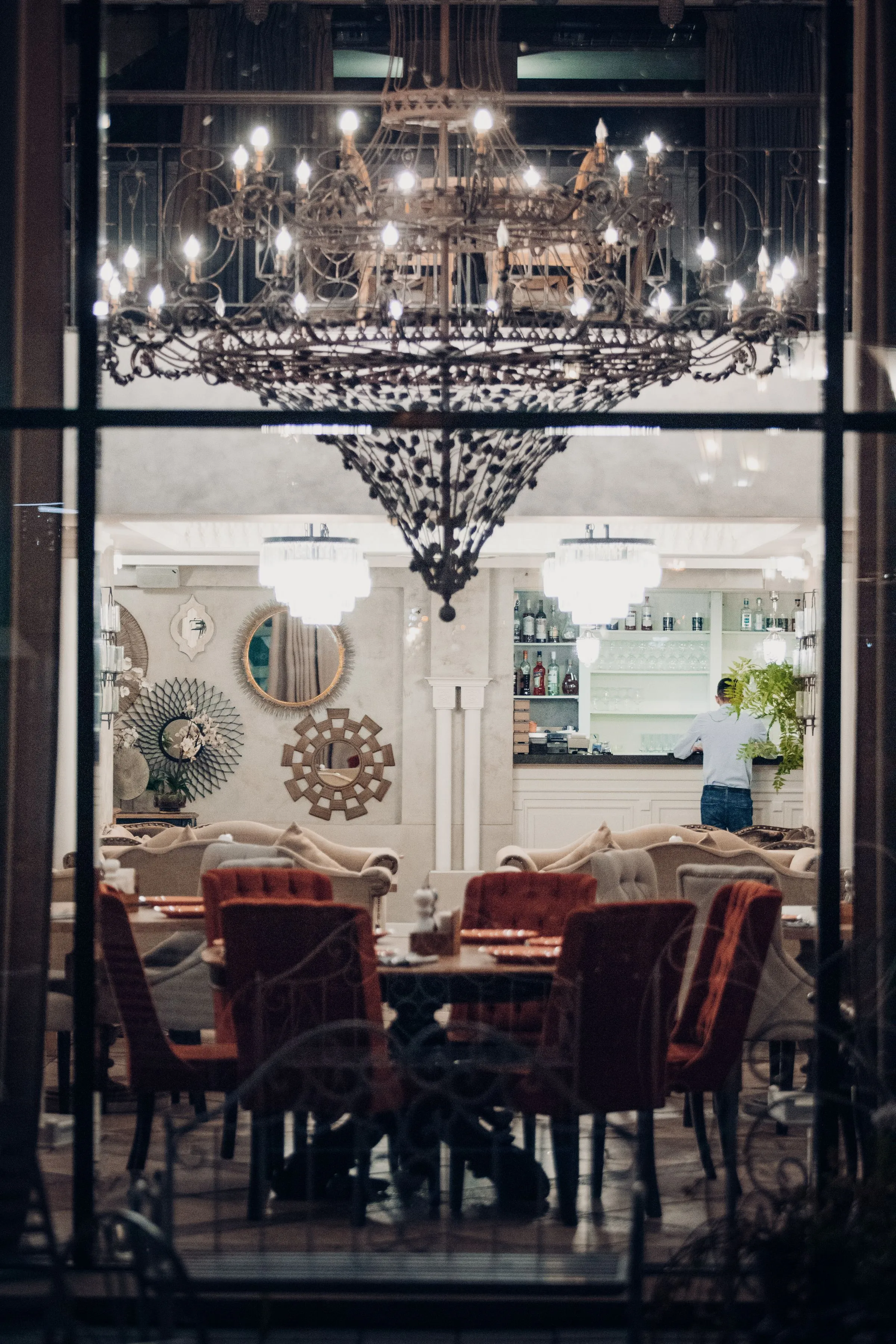 Restaurant at Hotel Marana seen through a window, with a chandelier above laid tables.