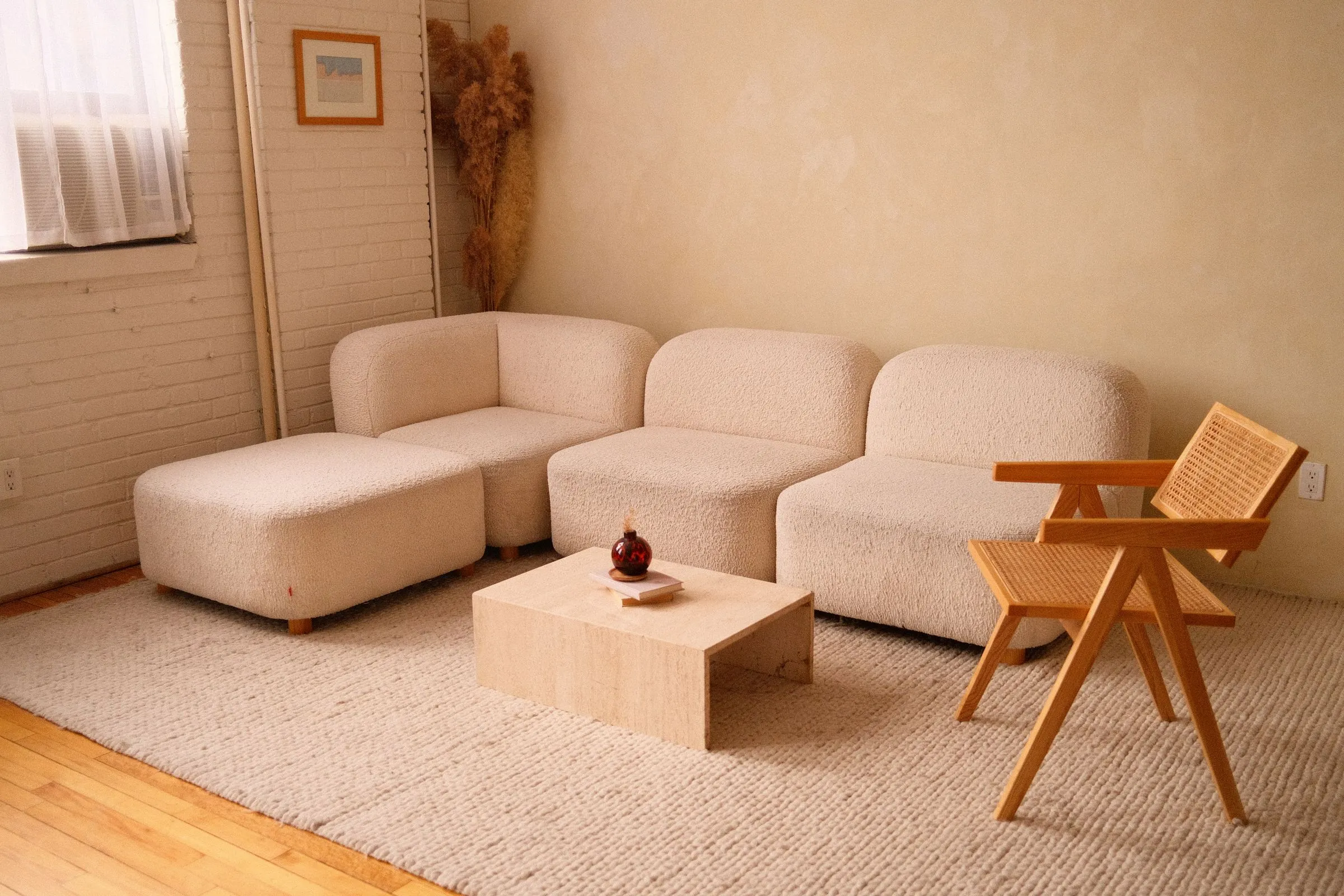 A calm living room with a low oatmeal modular sofa, a timber armchair and a bare plaster wall.