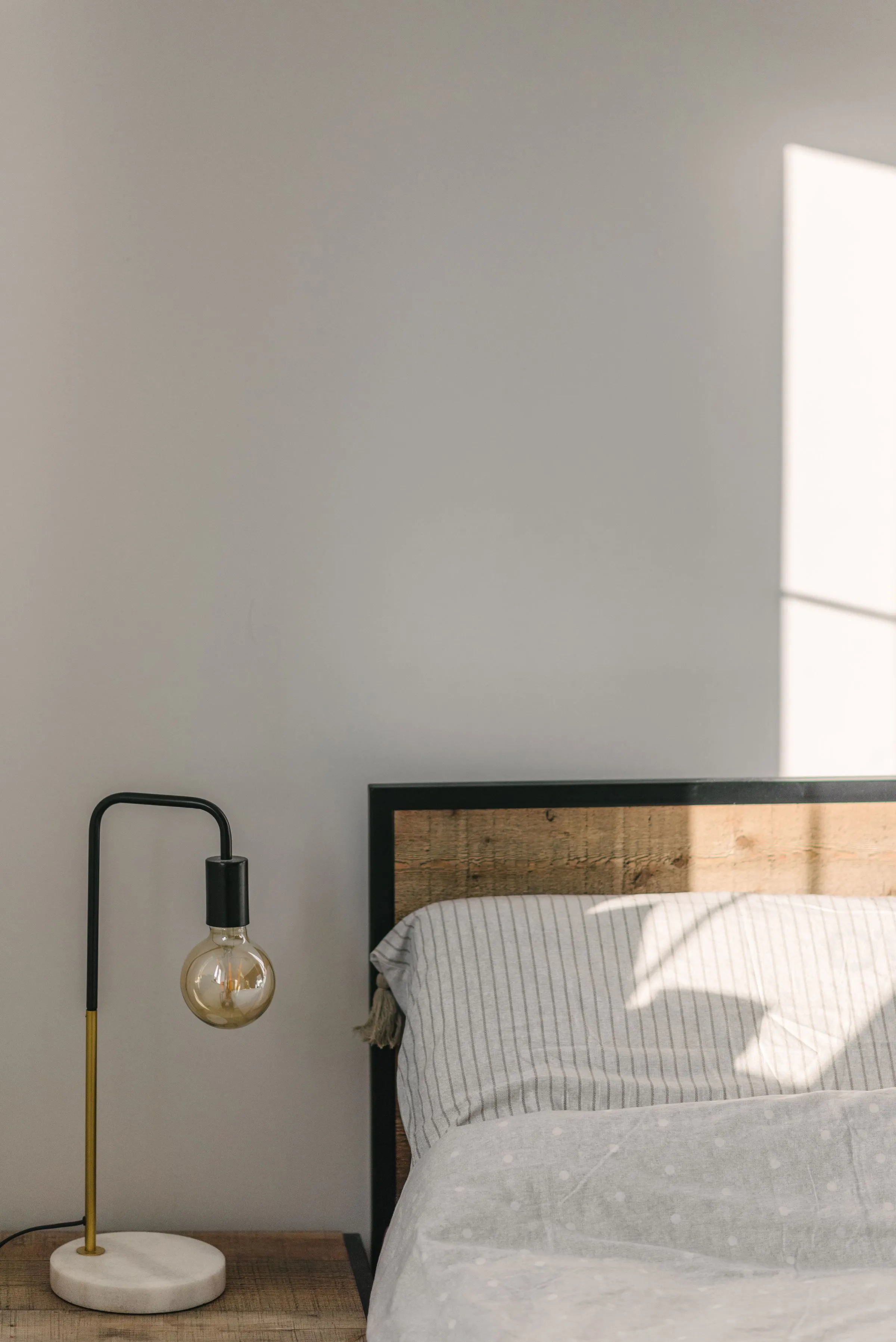 A bed with a crumpled blanket beside a timber table, with shadow across the wall.
