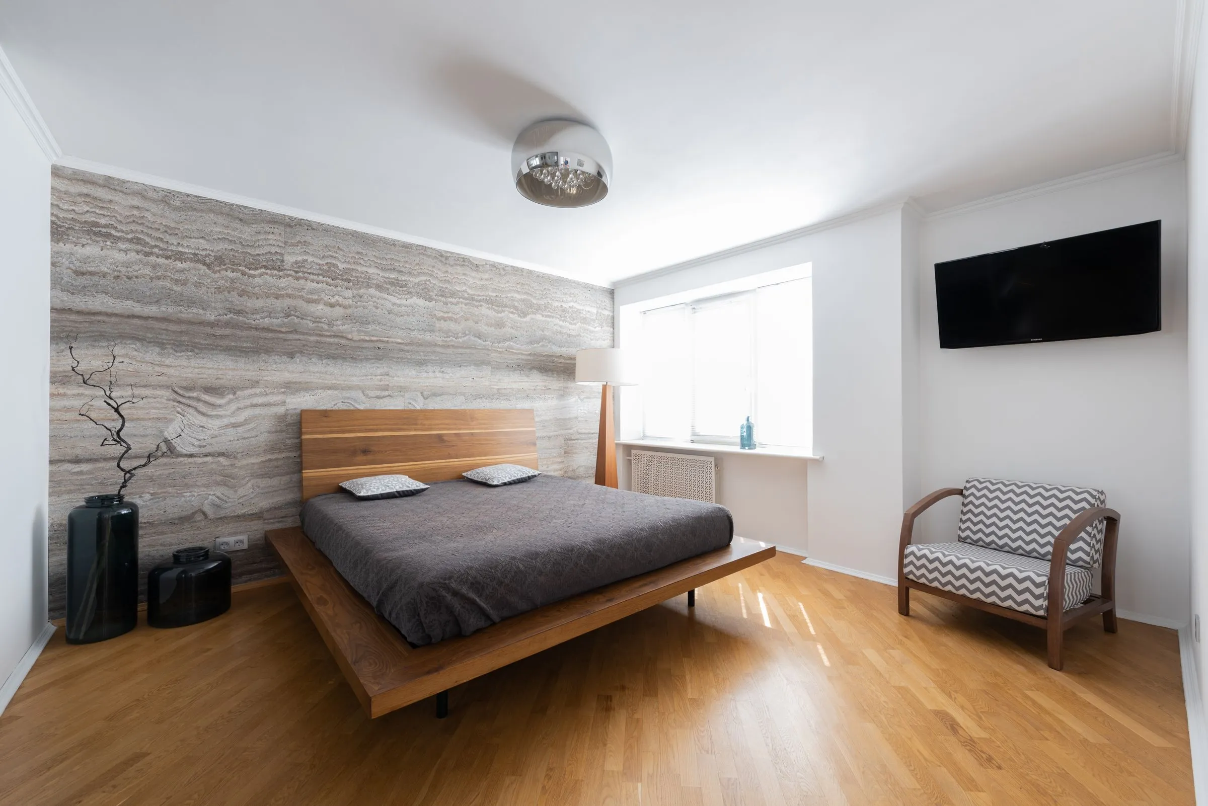 A light apartment interior with a bed, an armchair and a lamp beside the window.