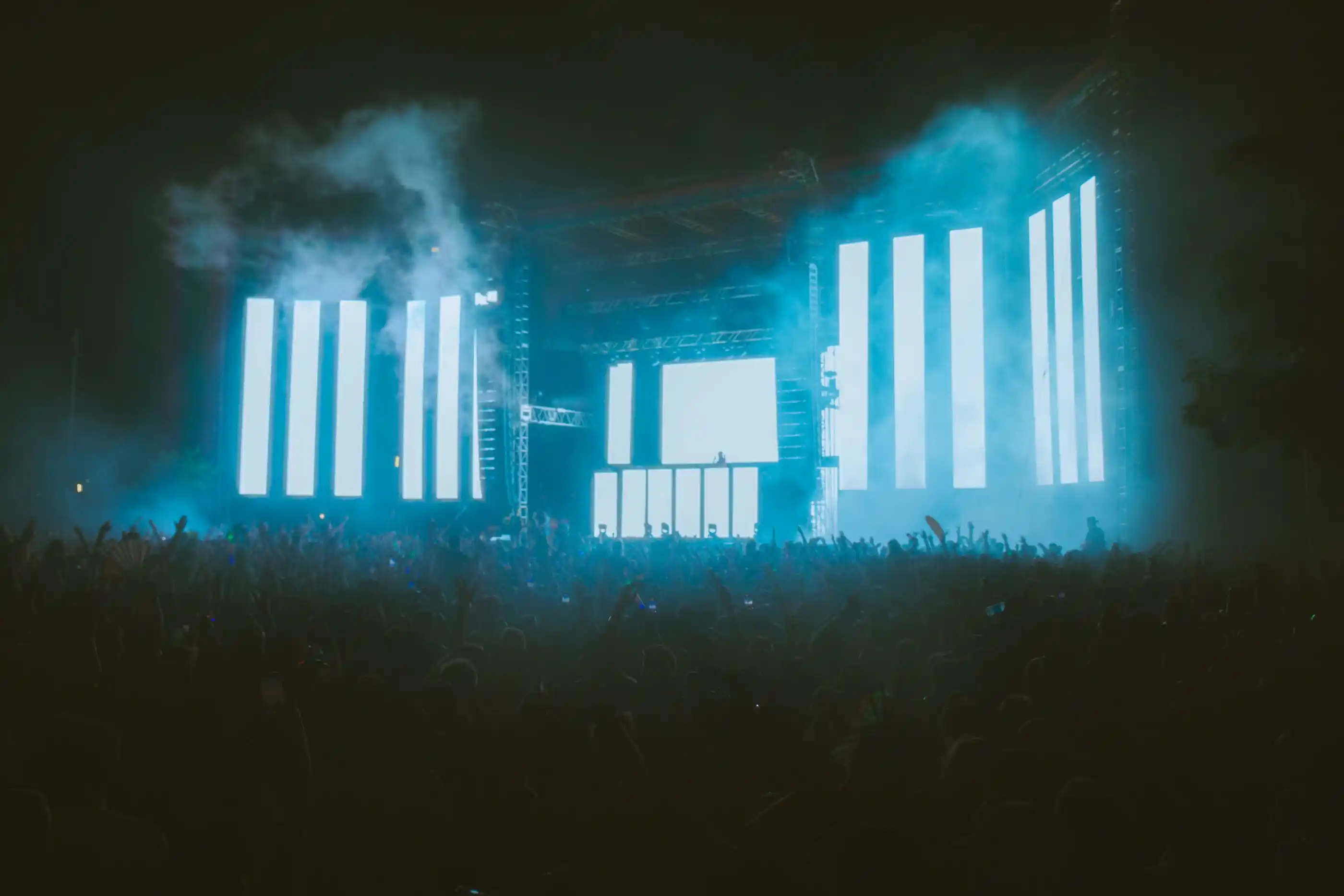 A festival crowd silhouetted against tall LED columns in low fog