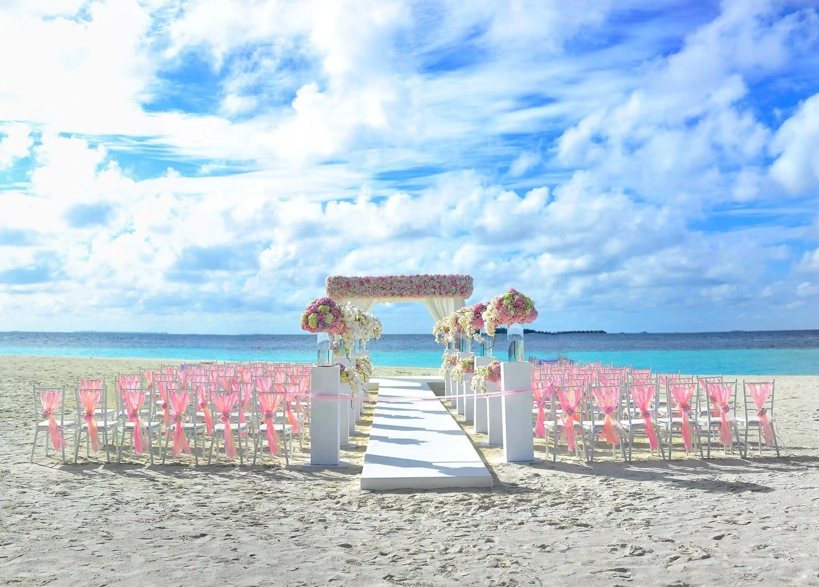 A ceremony set on white sand facing open water, chairs dressed in pale linen