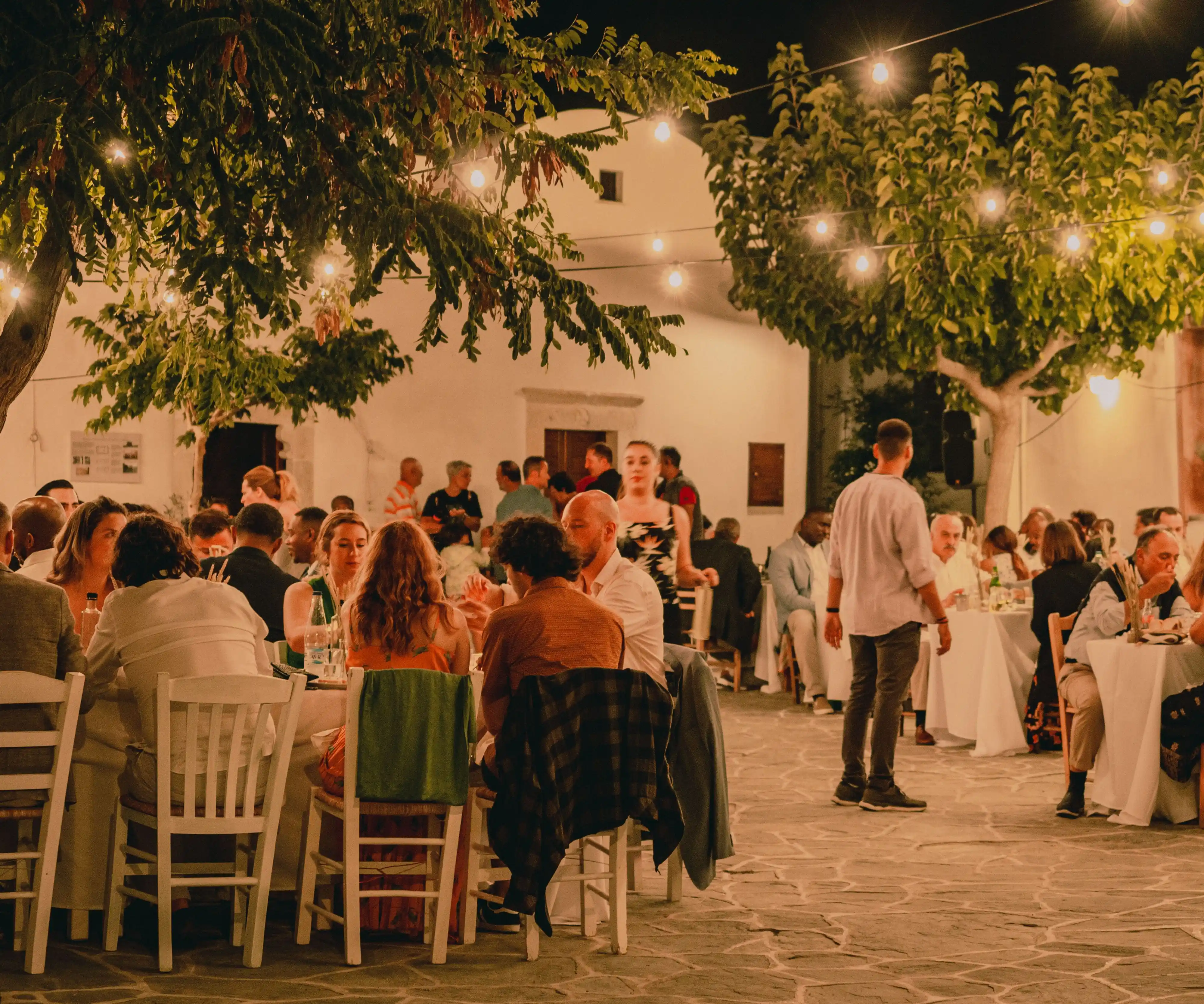 Guests dining at a long outdoor table under strings of warm bulbs after dark