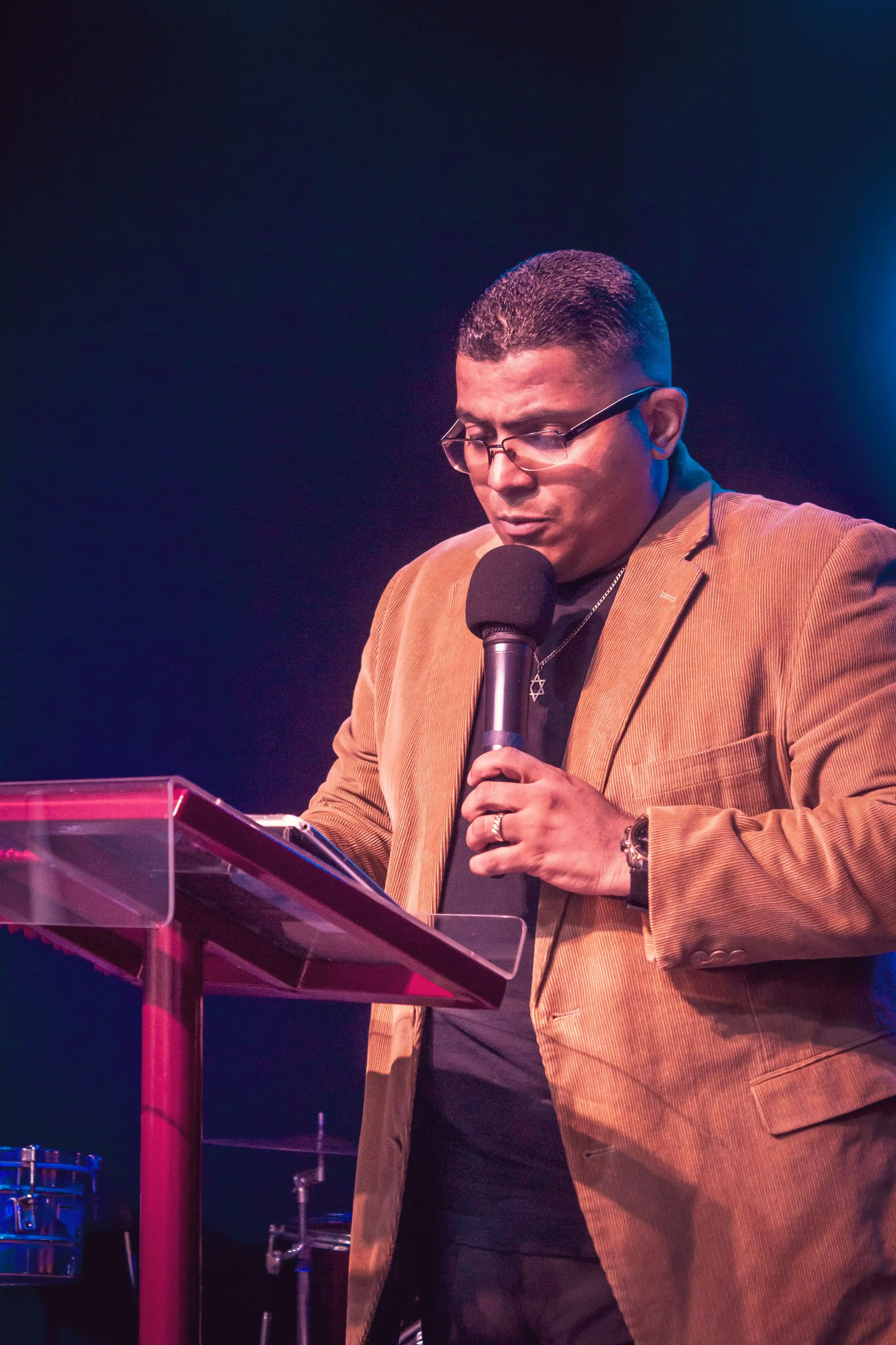 A speaker at a lectern under stage light, microphone in hand, mid-sentence