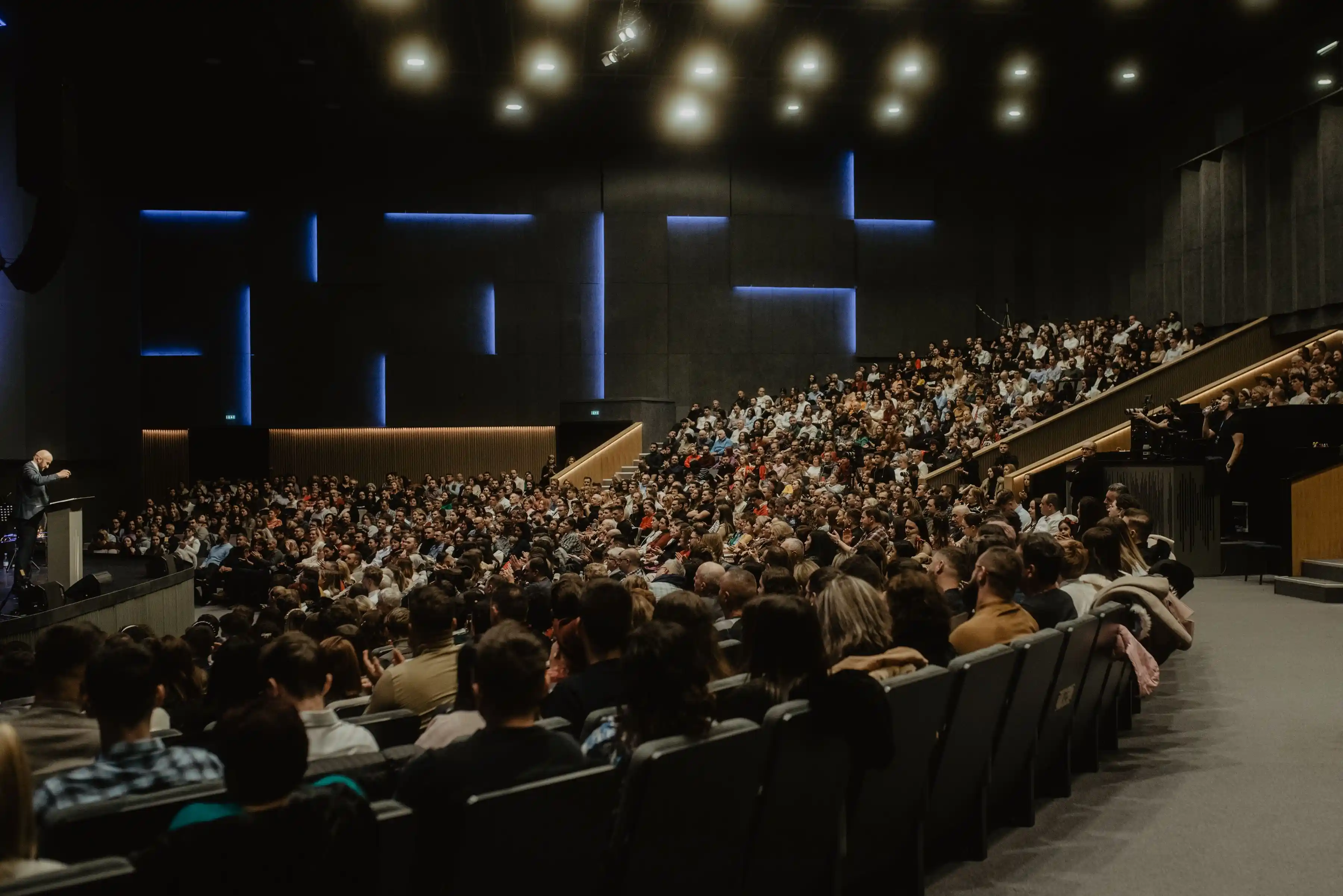 A large audience seated in a raked hall, attention on a distant stage