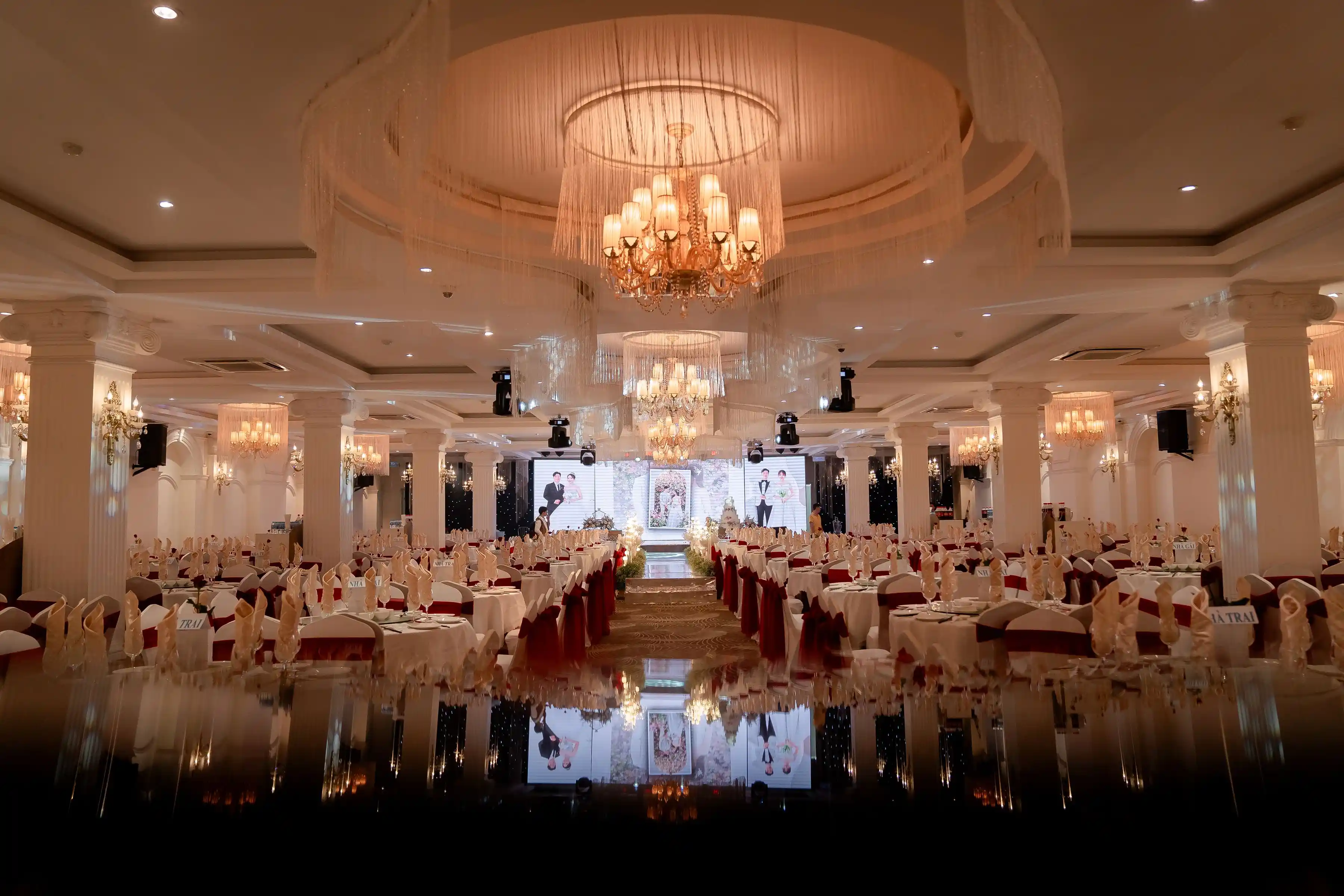 A grand ballroom dressed for a gala, long tables converging on a lit stage under a chandelier