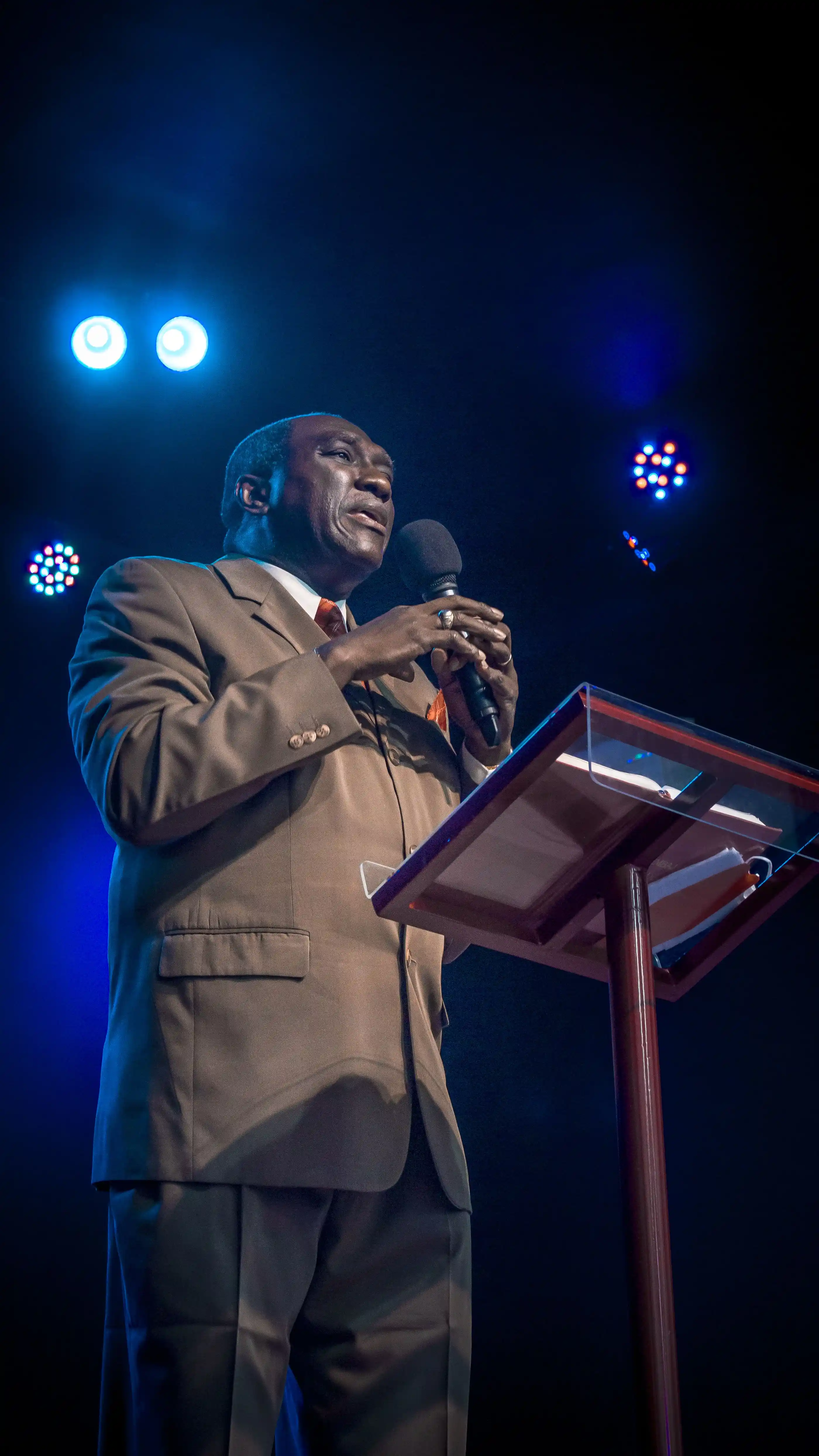 A speaker at a lectern under blue stage light, mid-address