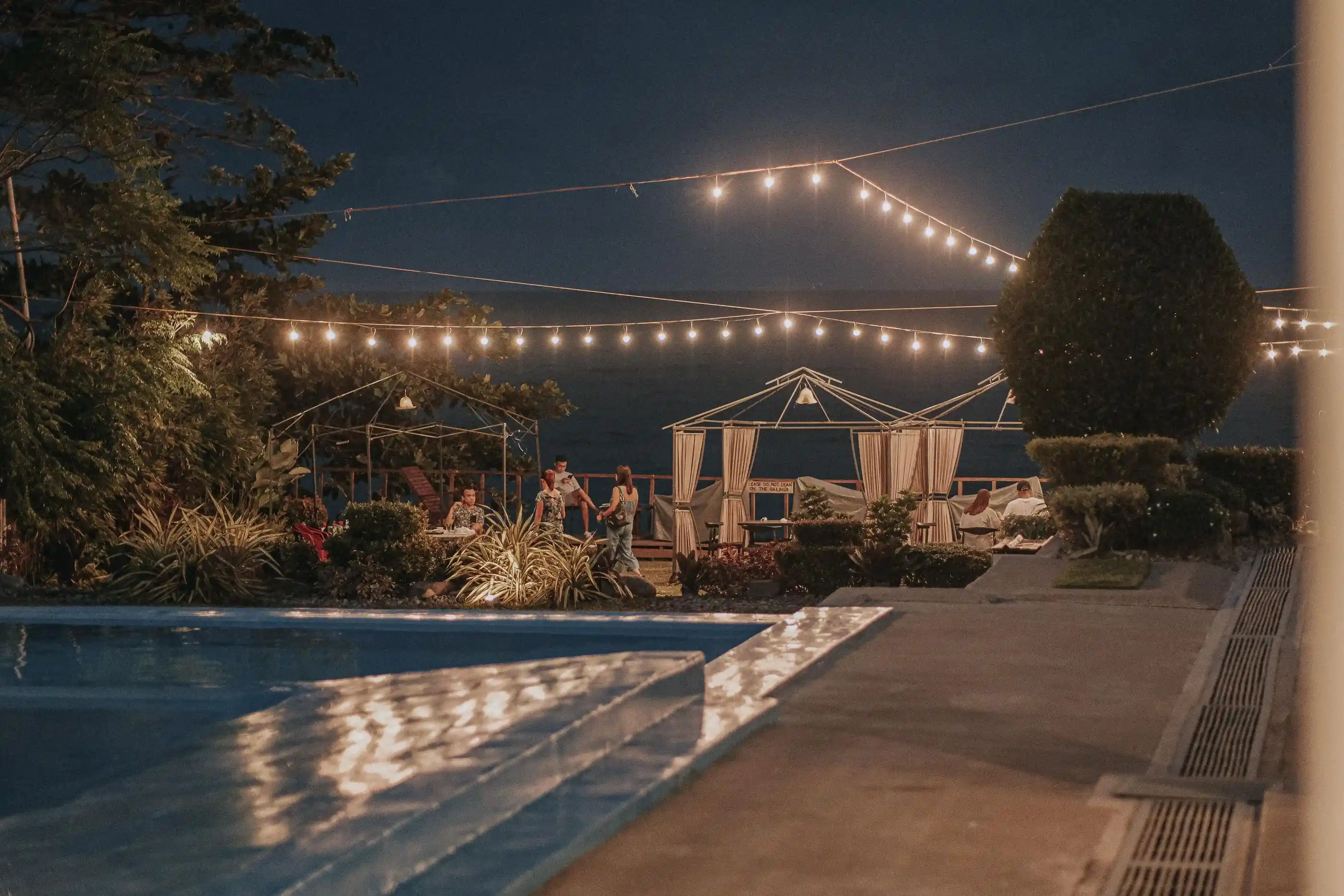 A poolside gathering at dusk under strings of bulbs