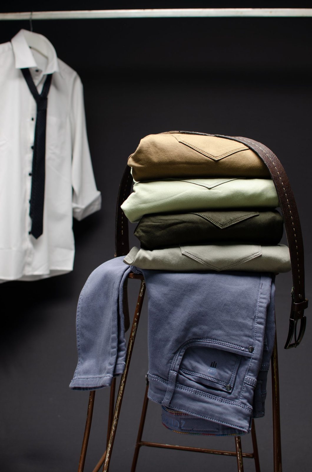 Dark editorial still life of folded knitwear and denim stacked on a stool