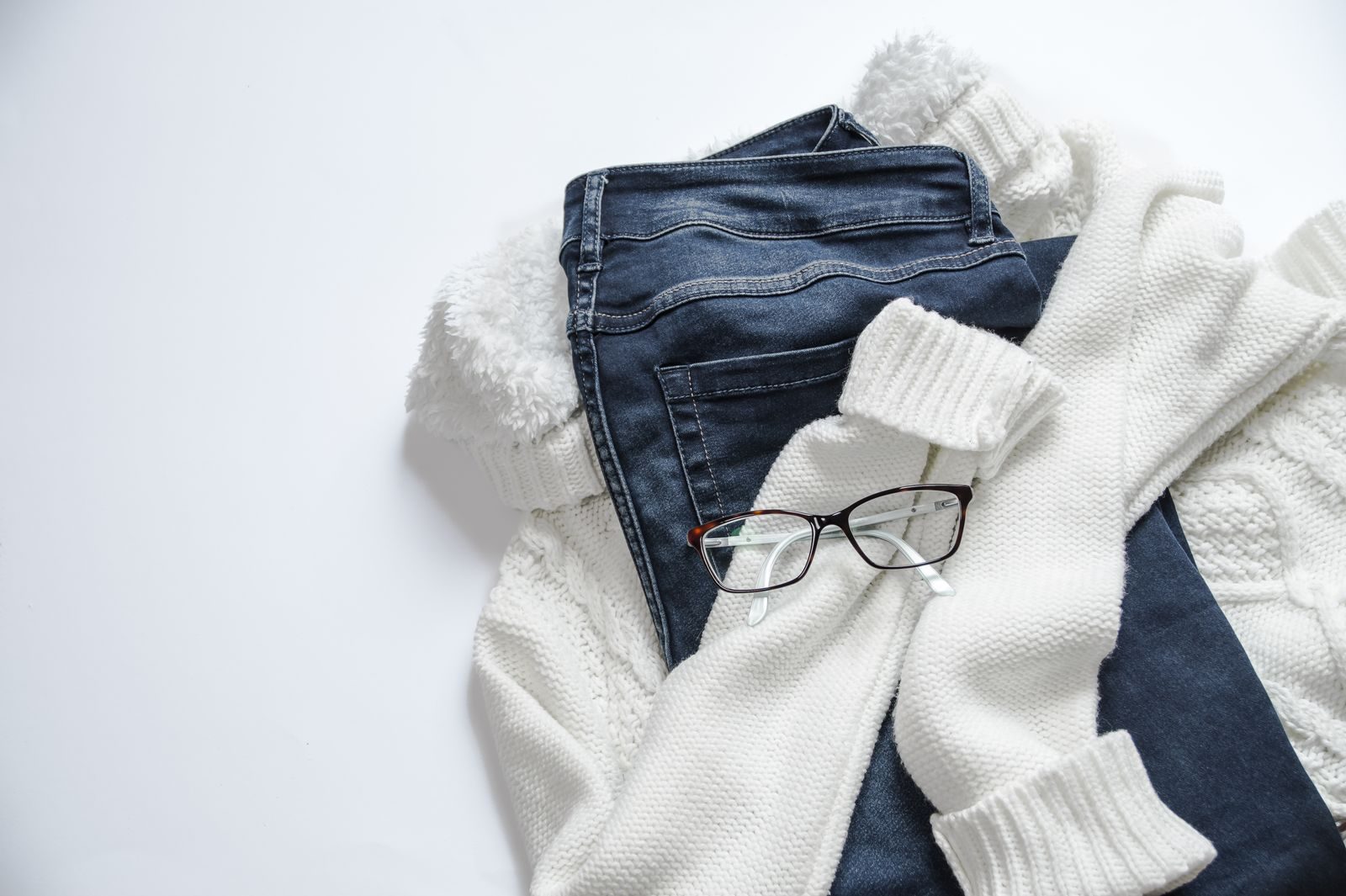 Flat lay of folded knitwear, denim and reading glasses arranged on a white surface
