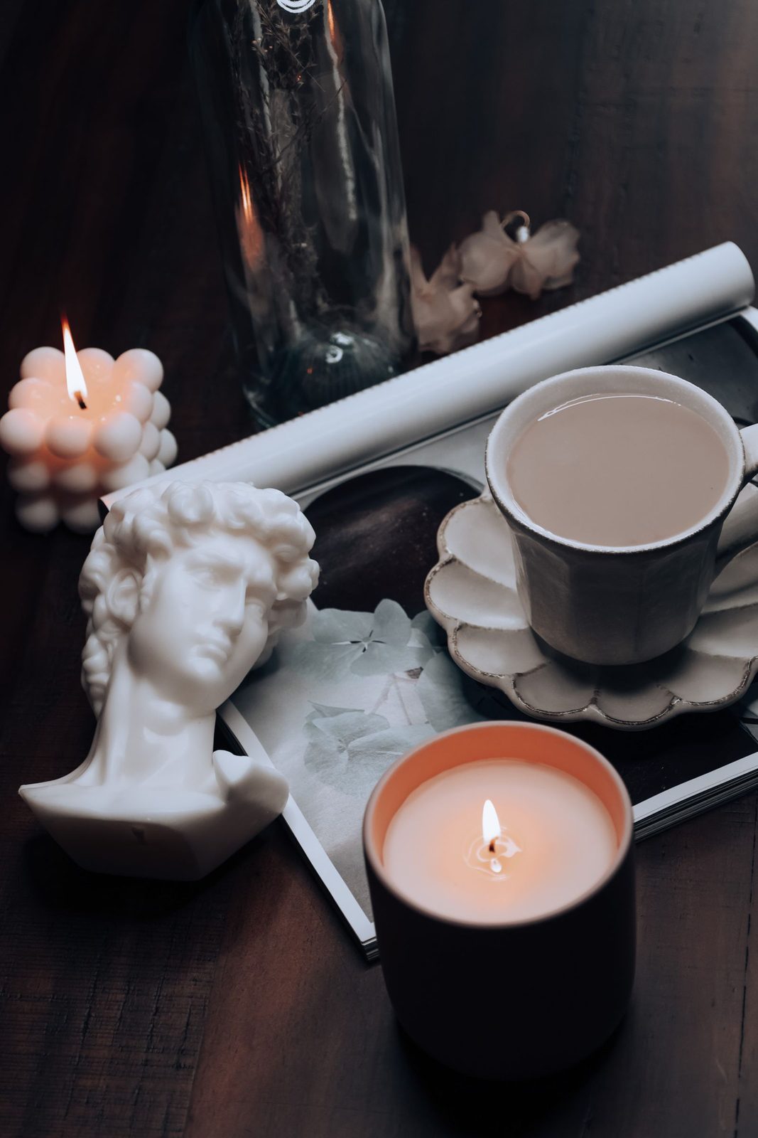 Lit Vesper candle beside a sculptural bust candle and ceramic cup on dark wood