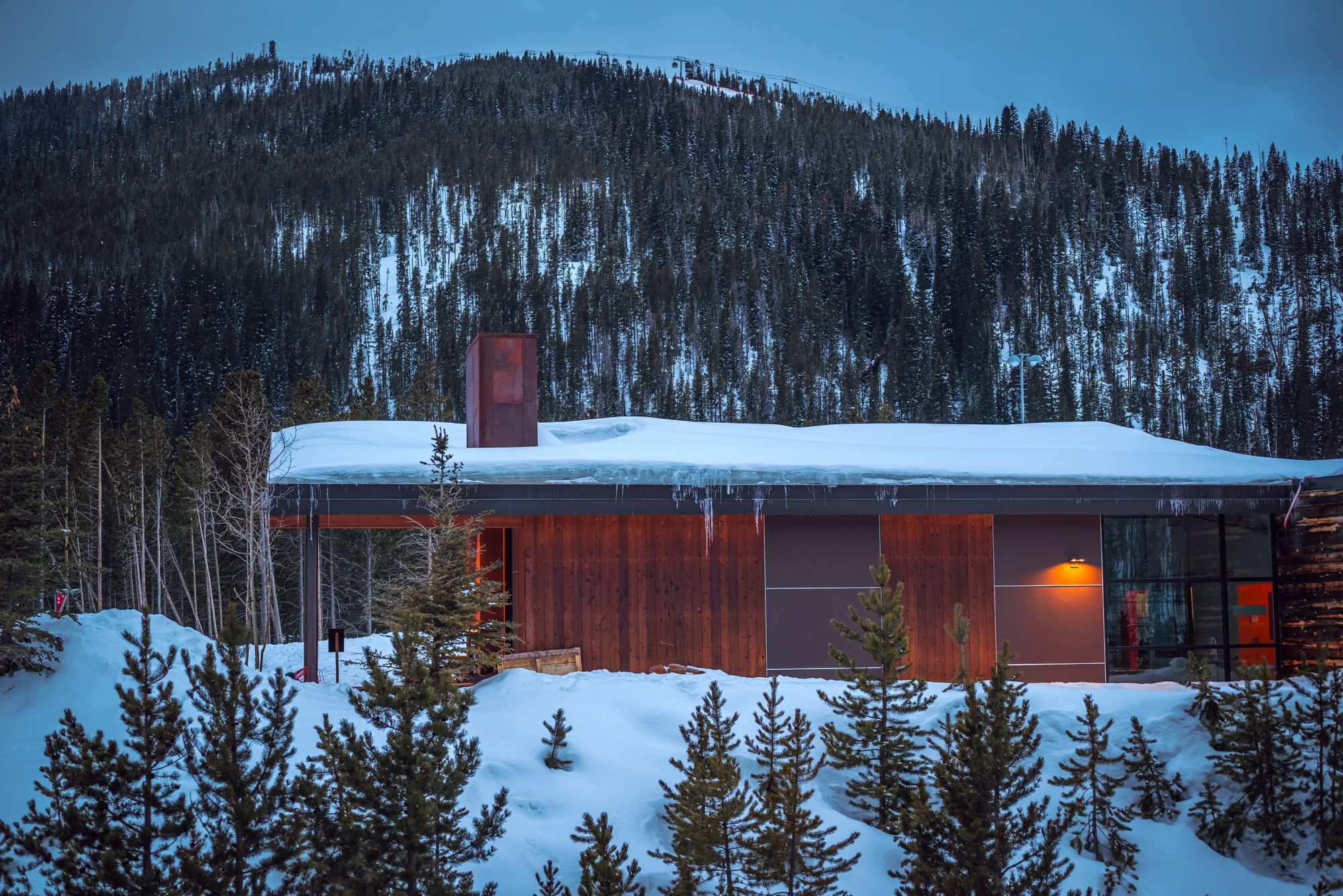 Exterior render of a blackened-timber highland lodge glowing at dusk against a snow-dusted mountainside