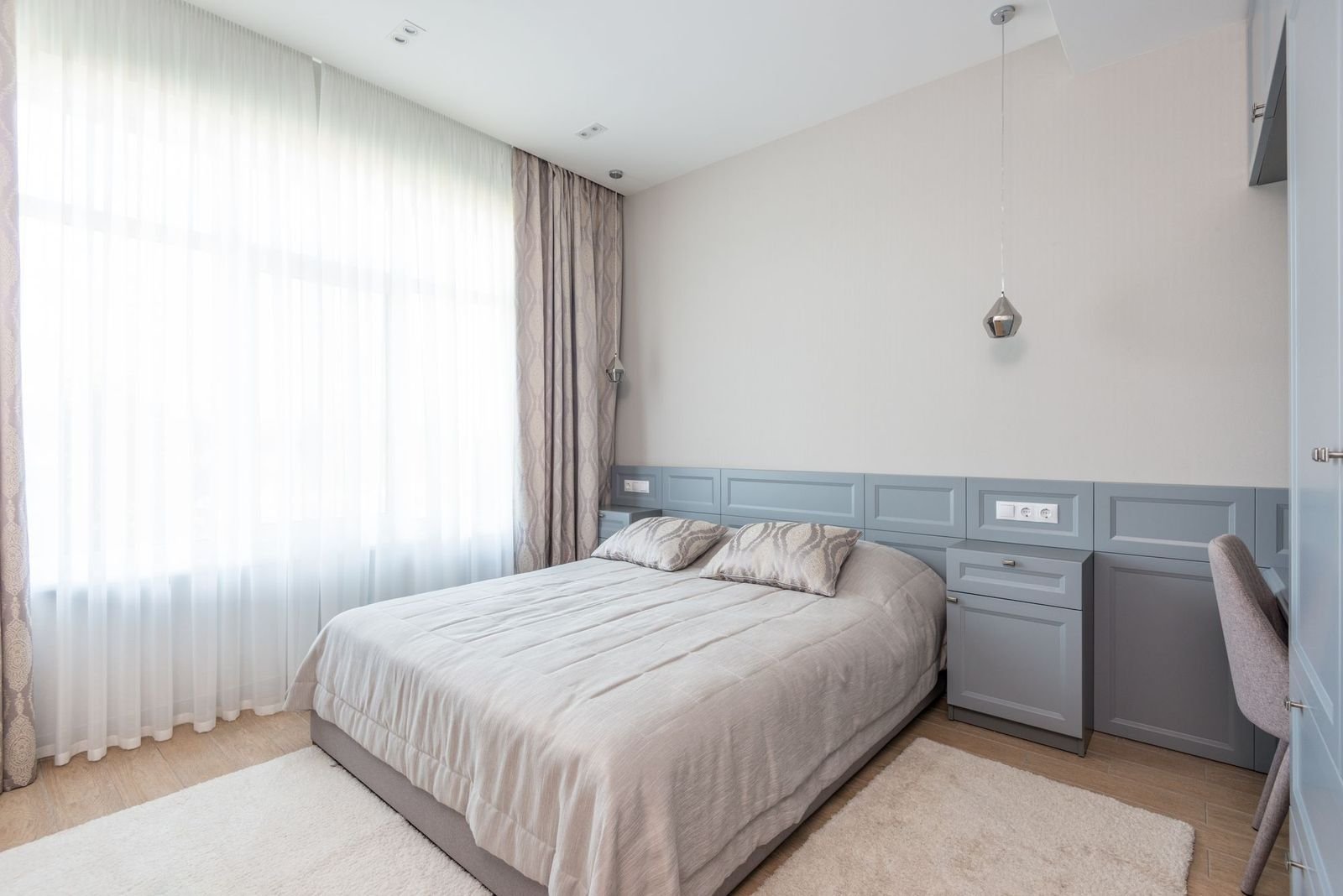 A hotel bedroom in calm neutrals, bed made flat, curtains drawn back