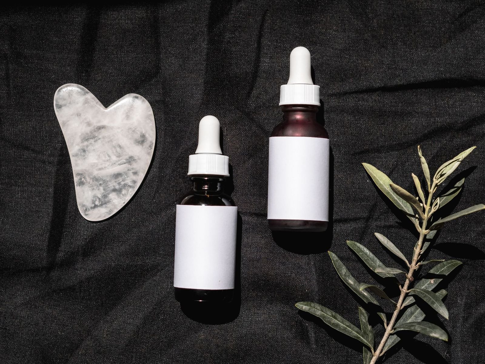 Skincare bottles and a gua sha arranged on dark folded fabric