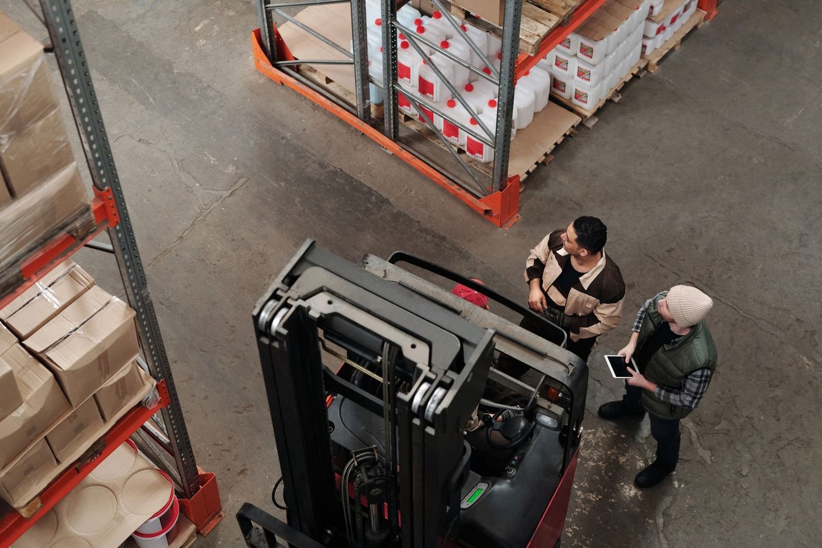 Two warehouse workers talking beside a forklift, viewed from a gantry