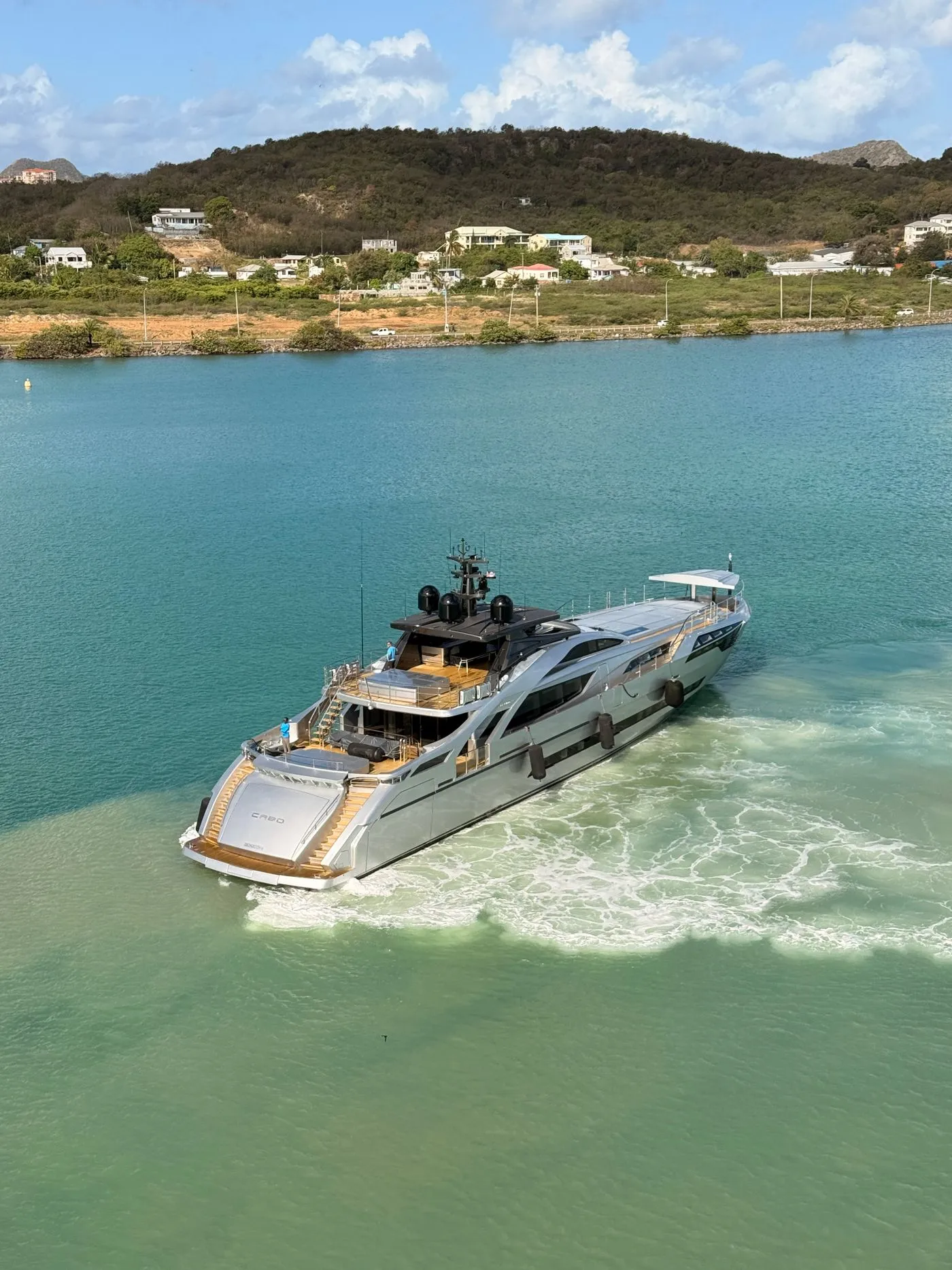 A luxury motor yacht cruising turquoise water below a green Mediterranean headland
