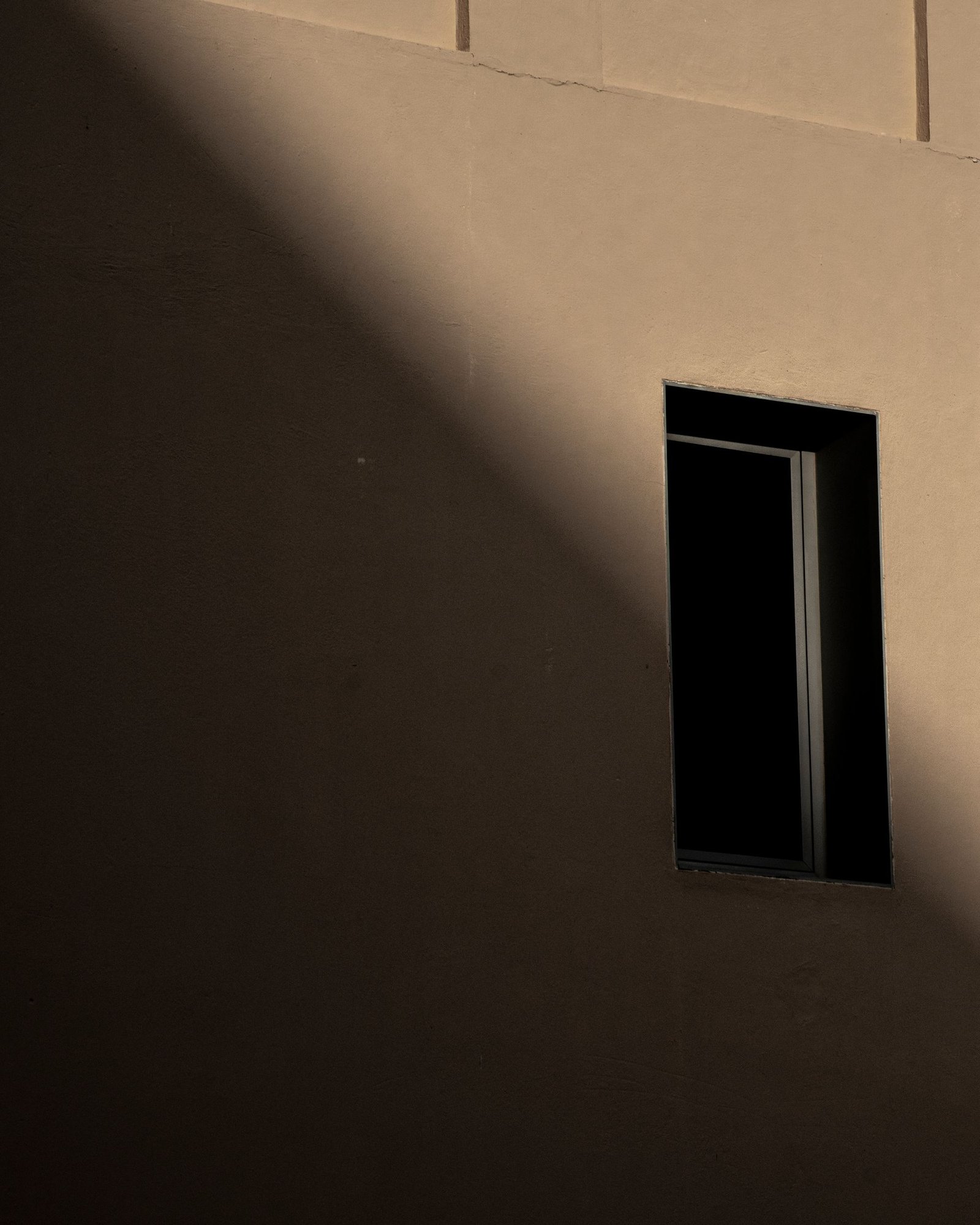 Sunlit plaster wall with a small deeply recessed window casting a hard shadow